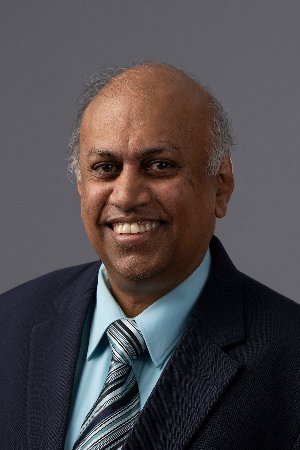 A smiling man wearing a navy blazer, light blue shirt, and striped tie against a gray background. The tone is professional and approachable.